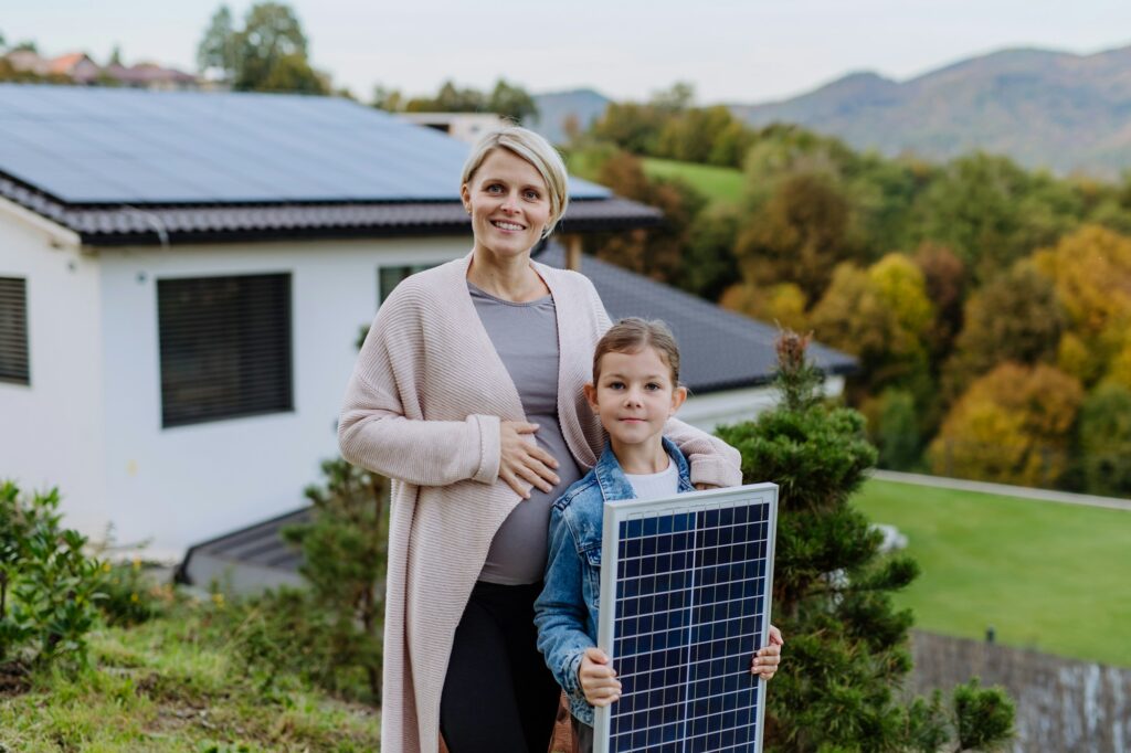 Mother with her little daughter near their house with solar panels. Alternative energy, saving